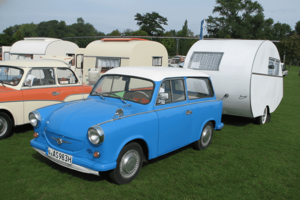 Blauer Trabant mit einem weißen Dübener-Ei-Wohnanhänger auf einer Wiese.