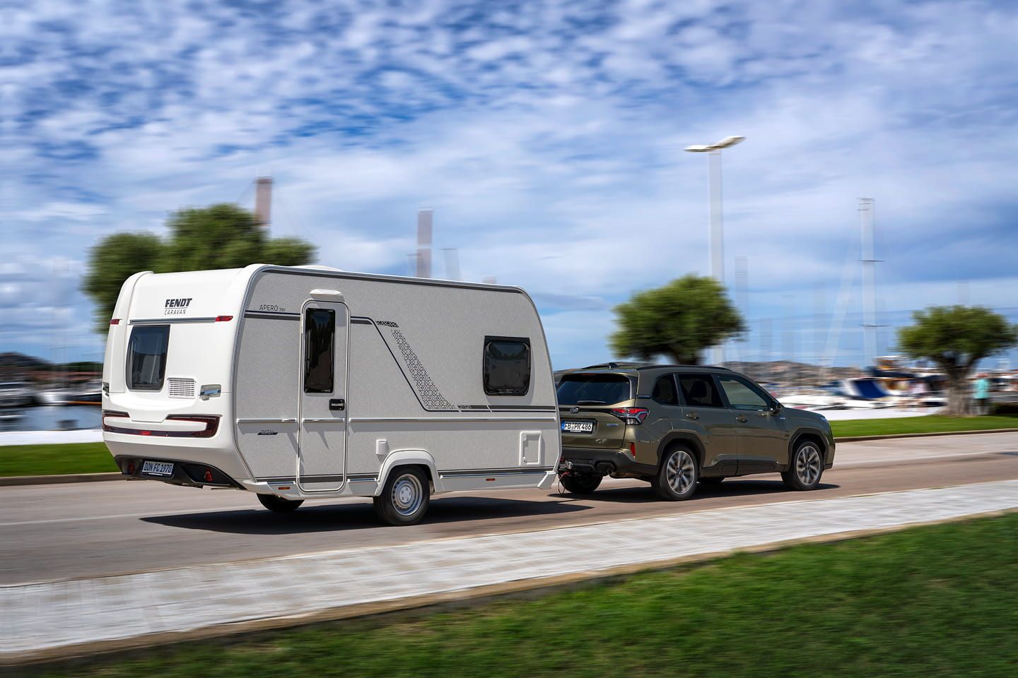 Wohnwagen wird von einem grünen SUV an einer Küstenstraße mit Hafen im Hintergrund gezogen