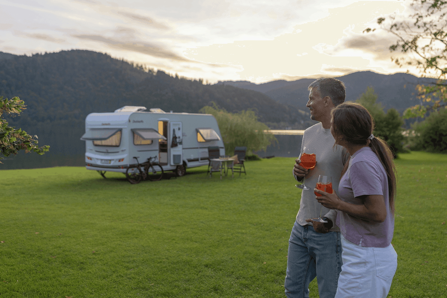 Paar mit Getränken steht abends auf einer Wiese am See, dahinter ein Wohnwagen mit Fahrrad und Campingmöbeln