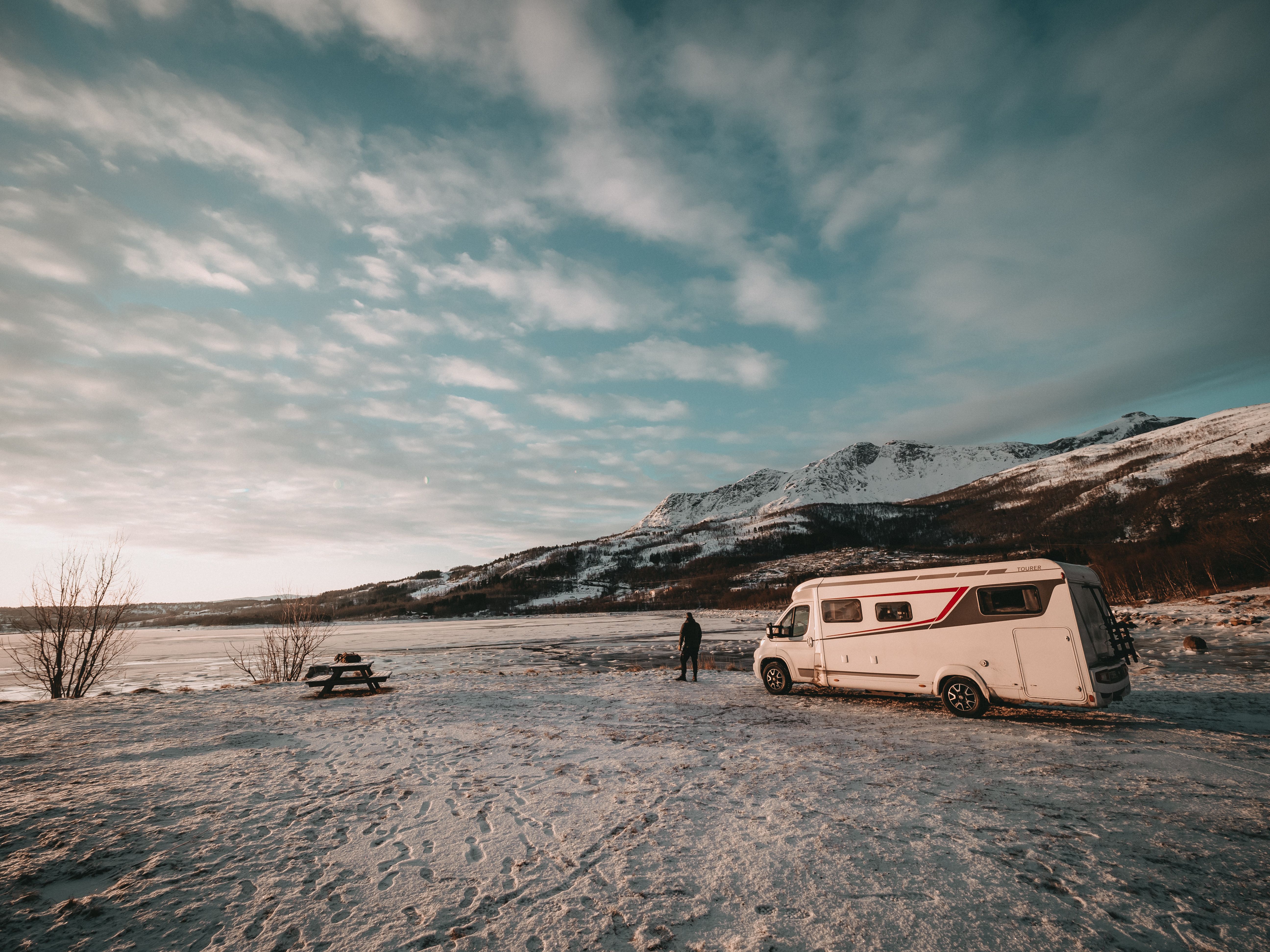 Wohnmobil an einem zugefrorenen See bei Abendlicht, davor ein Mann und eine Picknickbank, dahinter verschneite Berge