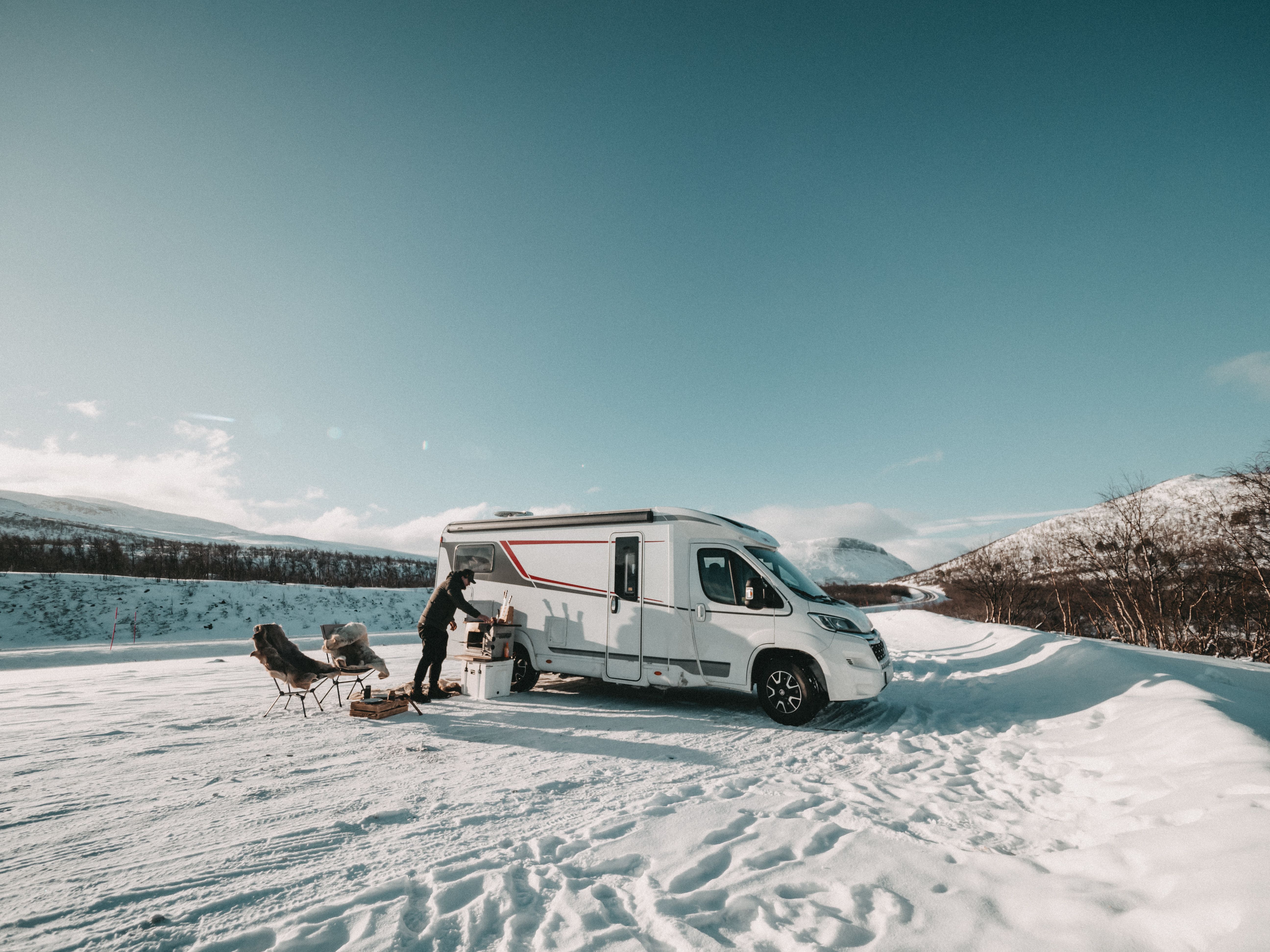 Weißes Wohnmobil im Schnee, davor Campingstühle und ein Mann beim Kochen unter blauem Himmel