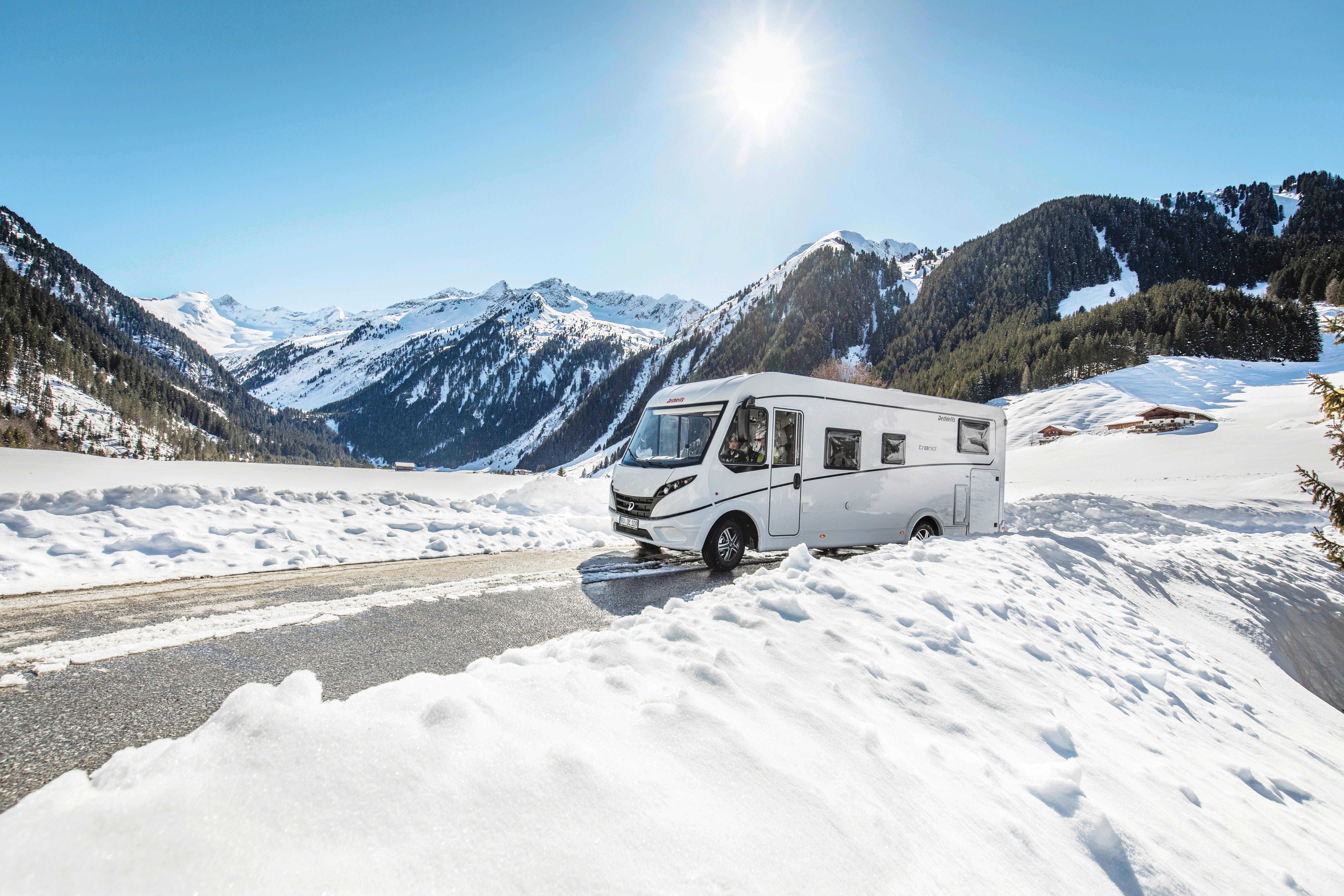 Weißes Wohnmobil auf einer Bergstraße zwischen Schneewällen, dahinter ein verschneites Alpenpanorama in der Sonne