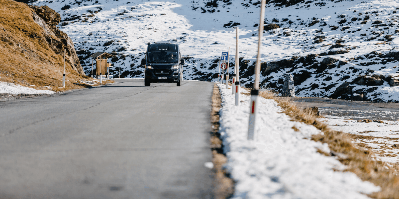 Ein Camper fährt bei Sonnenschein auf einer Straße. Am Straßenrand und im Hintergrund liegt Schnee.